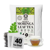 A white stand-up pouch of Akshit Premium Moringa Leaf Tea with a green leaf graphic on the label, shown alongside a stack of individual tea bags, a green moringa leaf sprig, and a glass cup of brewed tea on a white background.